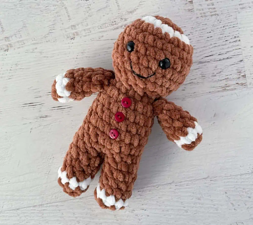 Crocheted plush gingerbread standing on a whitewashed table, features brown and white yarn and red buttons.