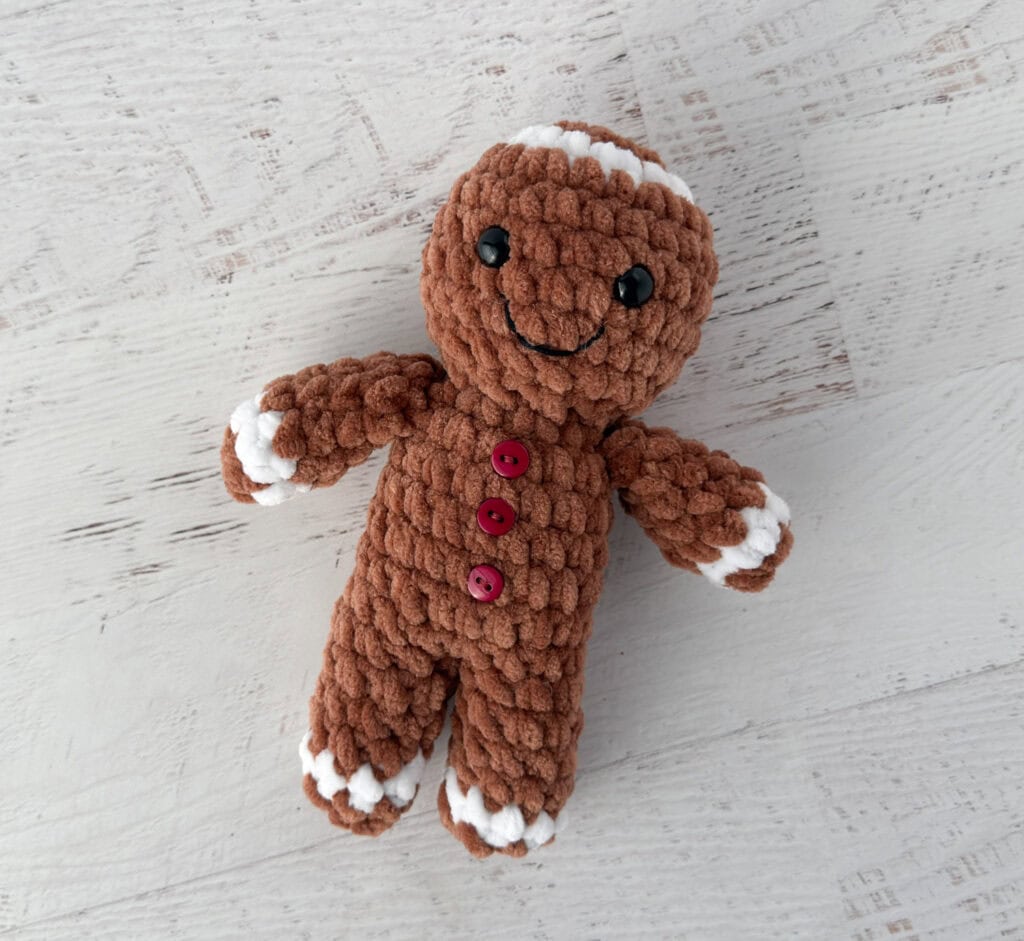 Crocheted plush gingerbread standing on a whitewashed table, features brown and white yarn and red buttons.