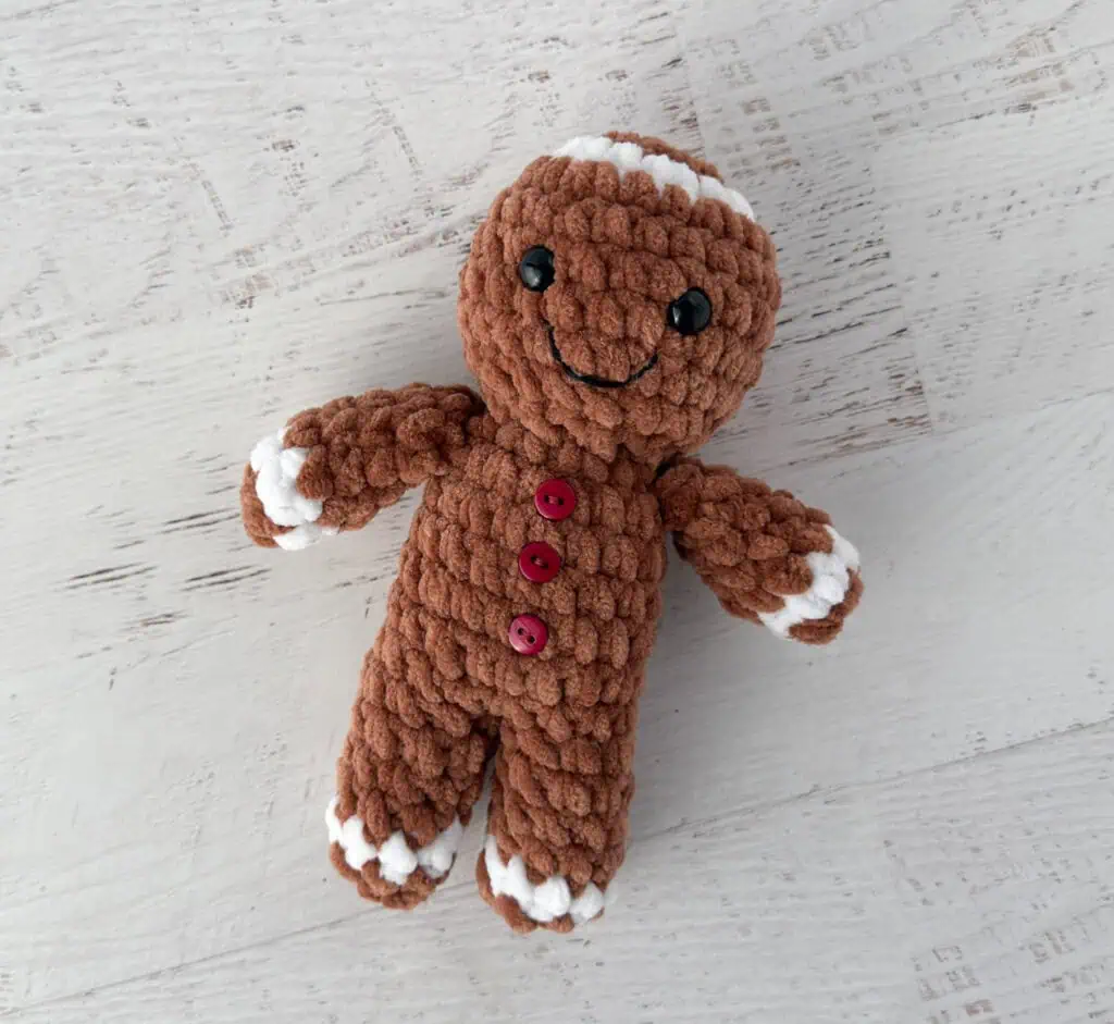 Crocheted plush gingerbread standing on a whitewashed table, features brown and white yarn and red buttons.