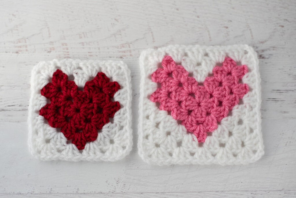 Two crochet heart squares, one red and one pink, on a white wooden surface