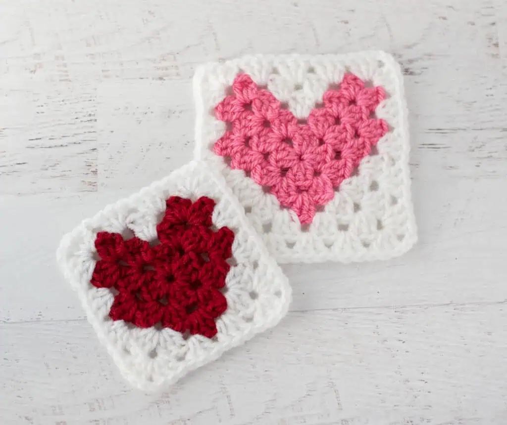 Two crochet heart squares, one red and one pink, on a white wooden surface