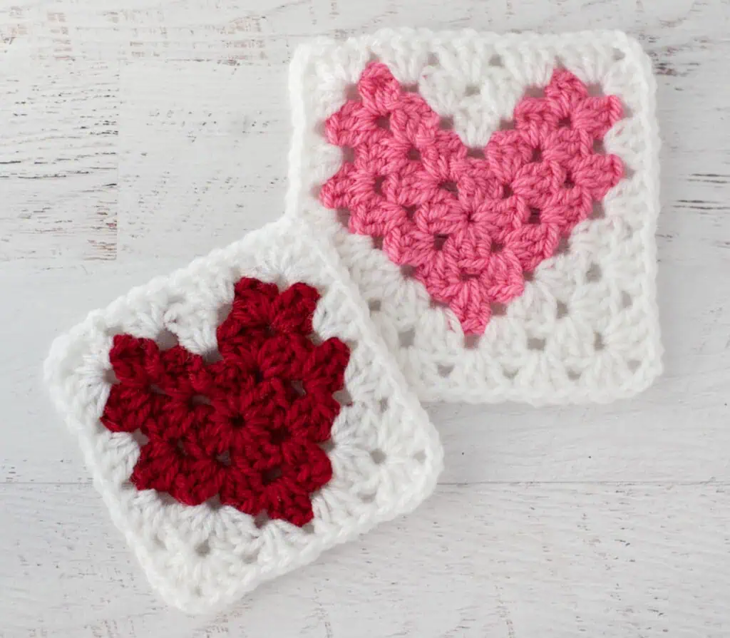 Two crochet heart squares, one red and one pink, on a white wooden surface