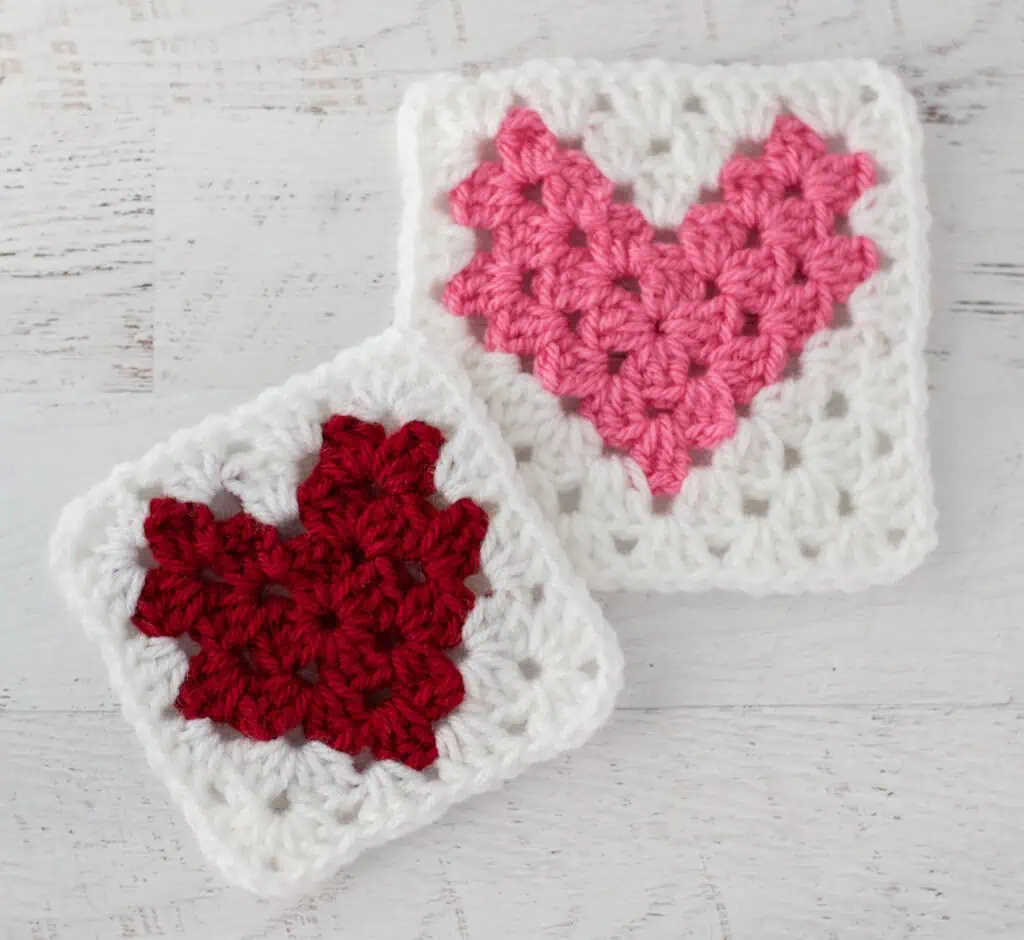 Two crochet heart squares, one red and one pink, on a white wooden surface