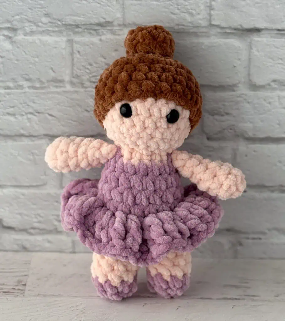 crochet ballerina with brown hair in purple dress standing in front of brick wall