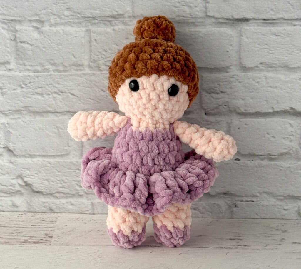 crochet ballerina with brown hair in purple dress standing in front of brick wall