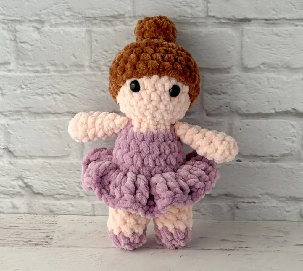 crochet ballerina with brown hair in purple dress standing in front of brick wall