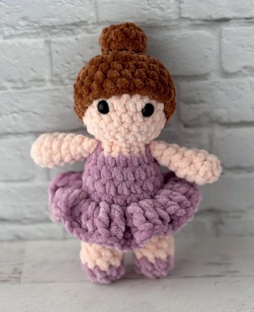 crochet ballerina with brown hair in purple dress standing in front of brick wall