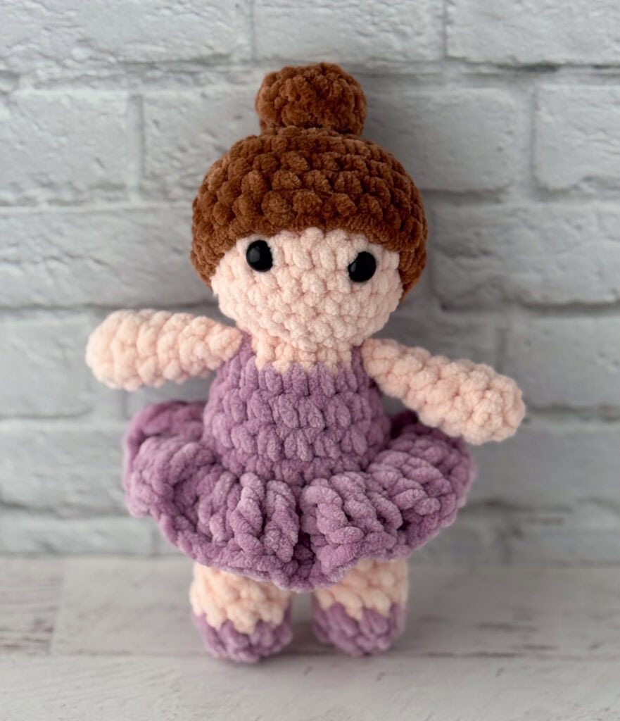 crochet ballerina with brown hair in purple dress standing in front of brick wall