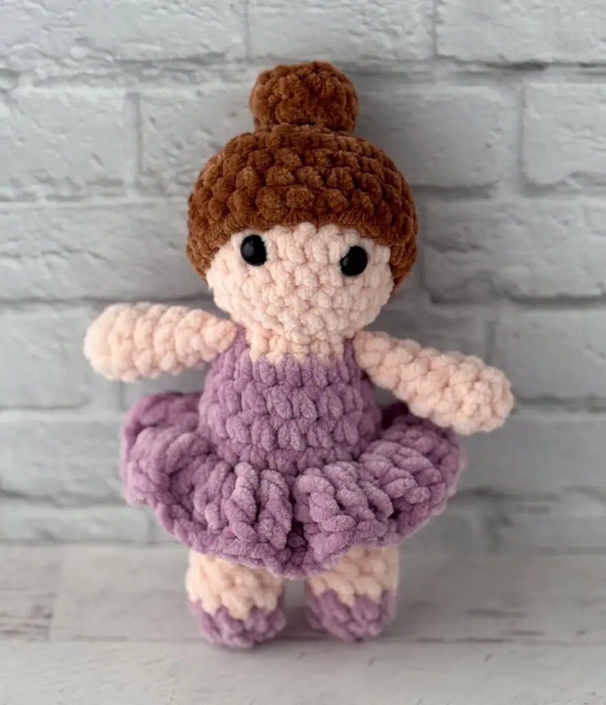 crochet ballerina with brown hair in purple dress standing in front of brick wall