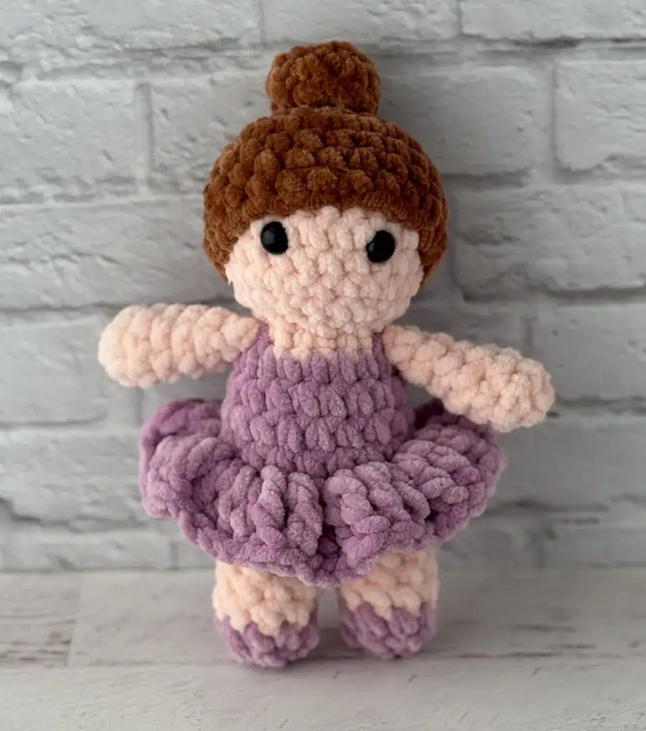 crochet ballerina with brown hair in purple dress standing in front of brick wall