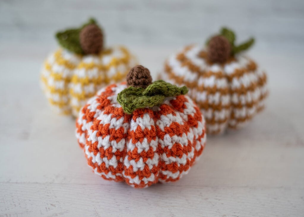 Set of 3 crochet houndstooth pumpkins in yellow, tan and orange yarn with white contrast yarn. Pumpkins have brown stems and green leaves.