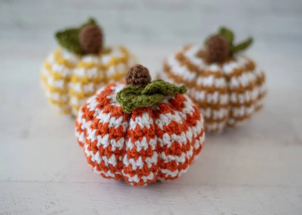 Set of 3 crochet houndstooth pumpkins in yellow, tan and orange yarn with white contrast yarn. Pumpkins have brown stems and green leaves.