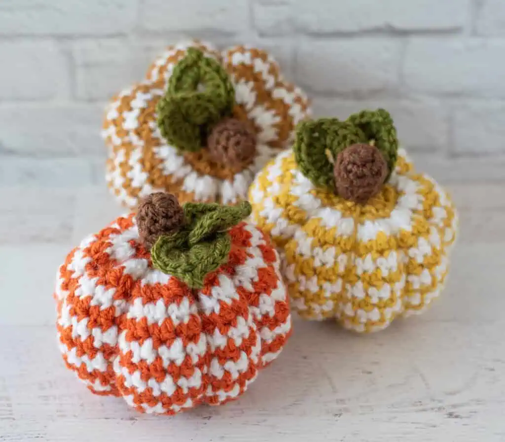 Set of 3 crochet houndstooth pumpkins in yellow, tan and orange yarn with white contrast yarn. Pumpkins have brown stems and green leaves.