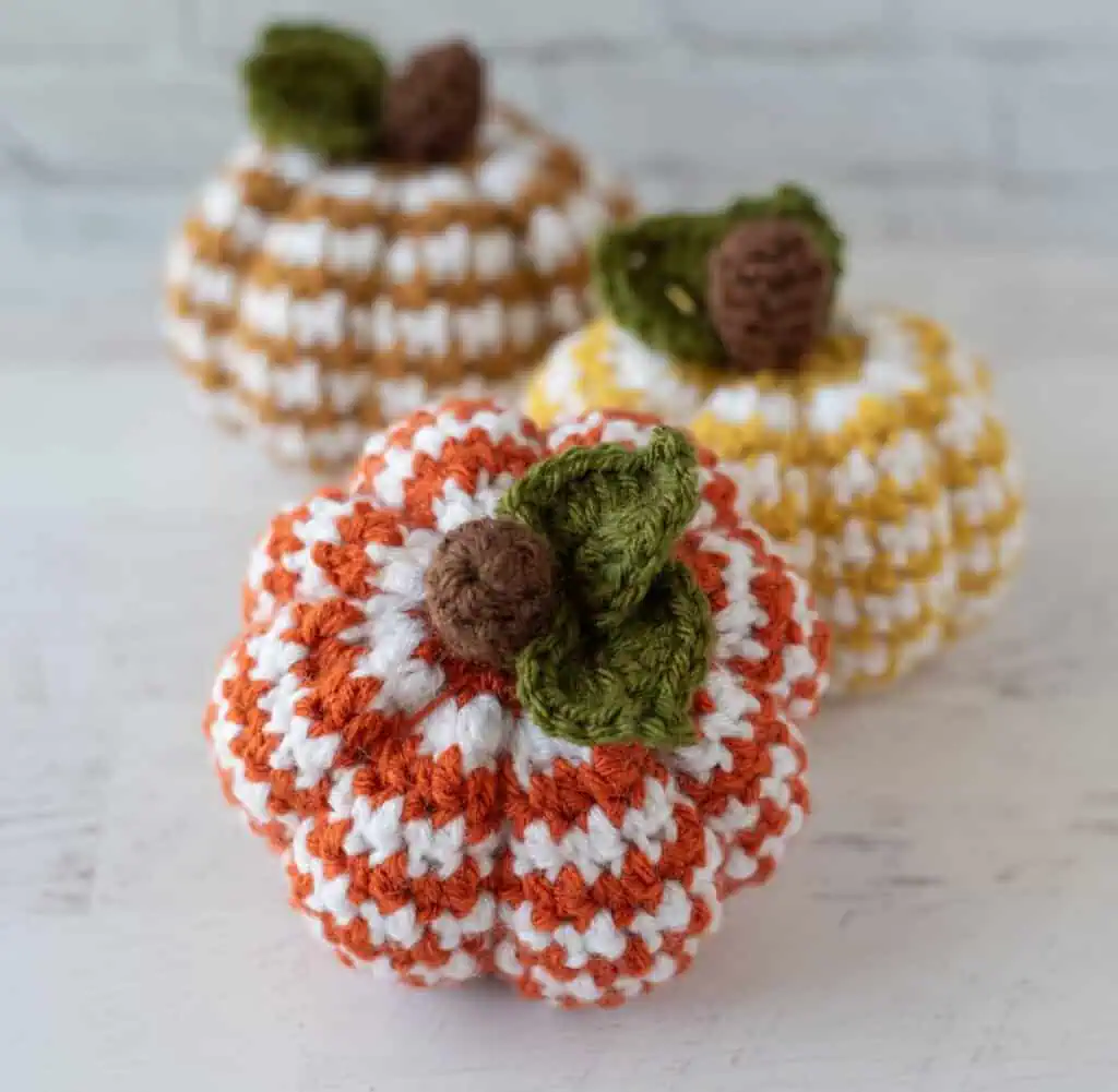 Set of 3 crochet houndstooth pumpkins in yellow, tan and orange yarn with white contrast yarn. Pumpkins have brown stems and green leaves.