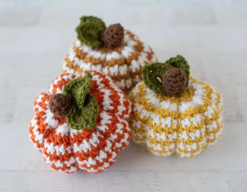 Set of 3 crochet houndstooth pumpkins in yellow, tan and orange yarn with white contrast yarn. Pumpkins have brown stems and green leaves.