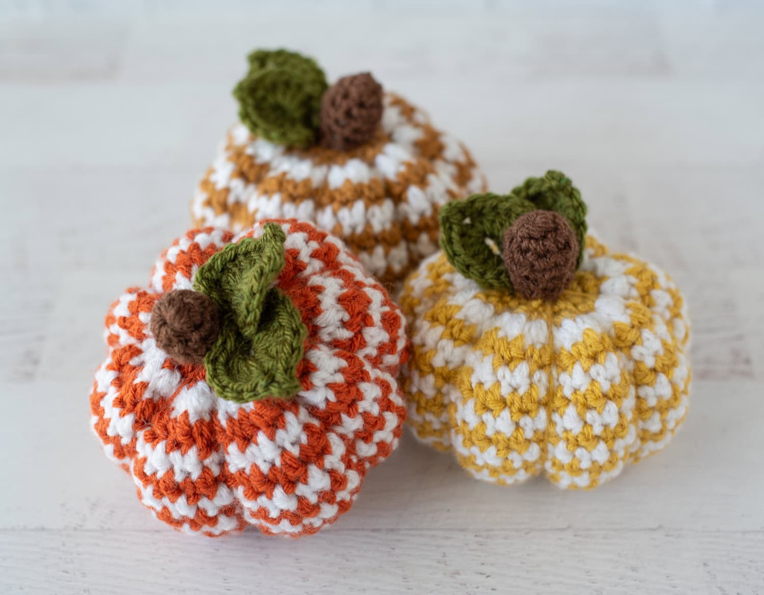 Set of 3 crochet houndstooth pumpkins in yellow, tan and orange yarn with white contrast yarn. Pumpkins have brown stems and green leaves.