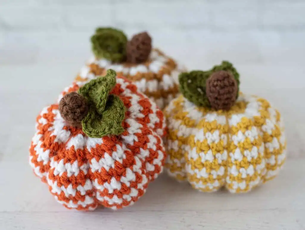 Set of 3 crochet houndstooth pumpkins in yellow, tan and orange yarn with white contrast yarn. Pumpkins have brown stems and green leaves. Orange pumpkin is propped against other pumpkins.