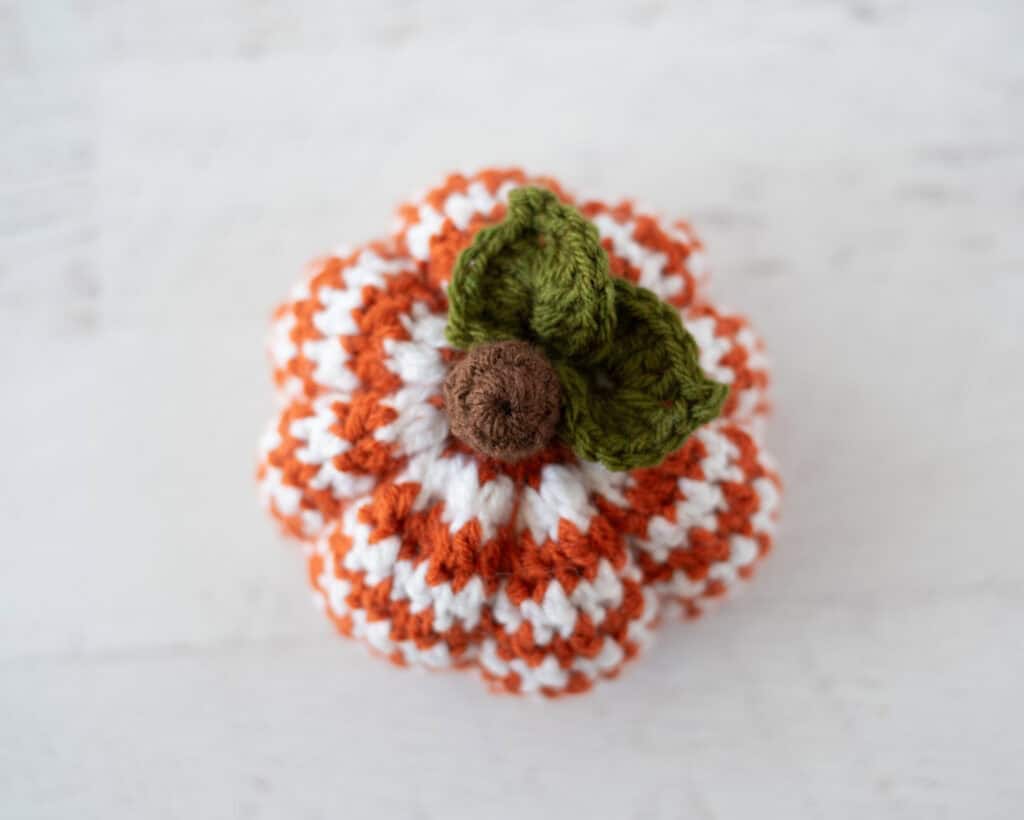 Top down view of crochet houndstooth patterned pumpkin in orange and white yarn with brown stem and green leaves
