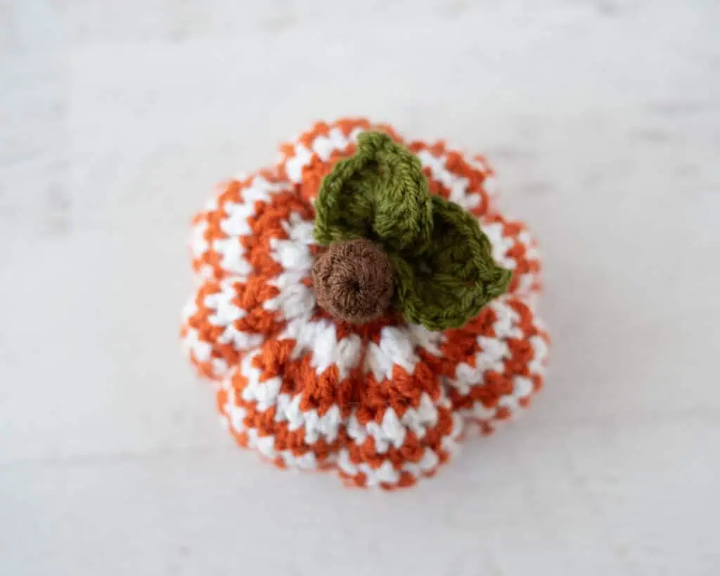 Top down view of crochet houndstooth patterned pumpkin in orange and white yarn with brown stem and green leaves