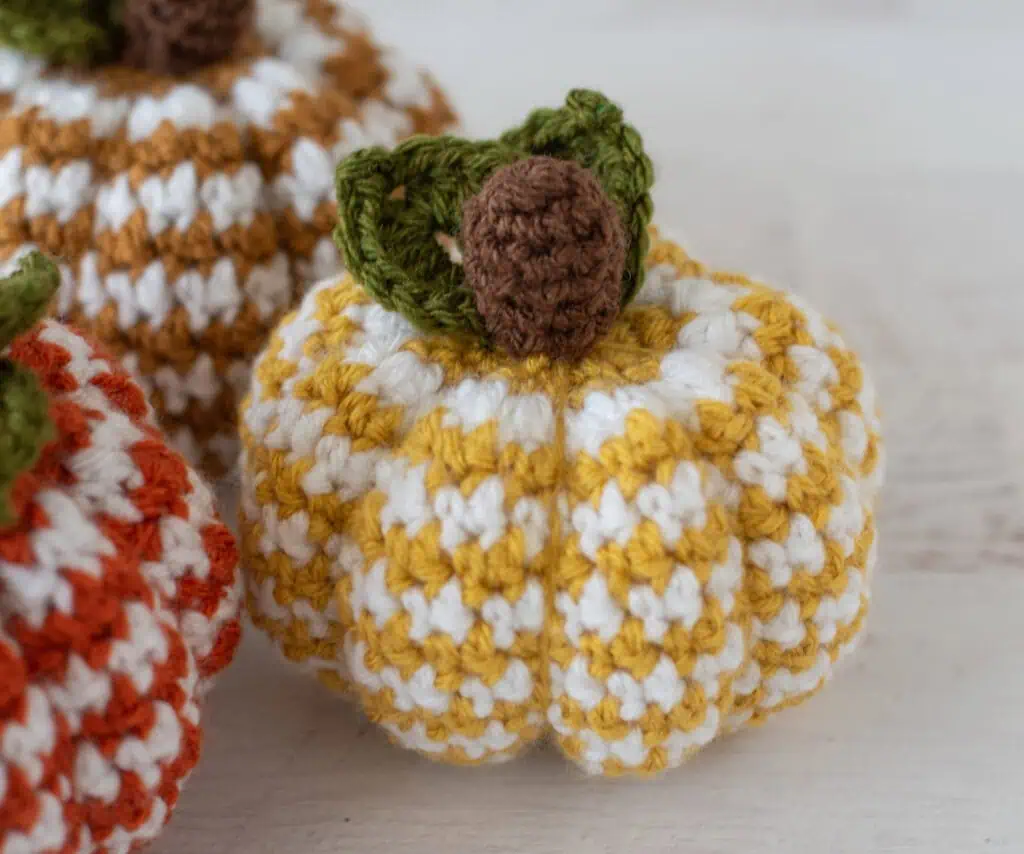 Set of 3 crochet houndstooth pumpkins in yellow, tan and orange yarn with white contrast yarn. Pumpkins have brown stems and green leaves. Focus is on yelllow pumpkin.