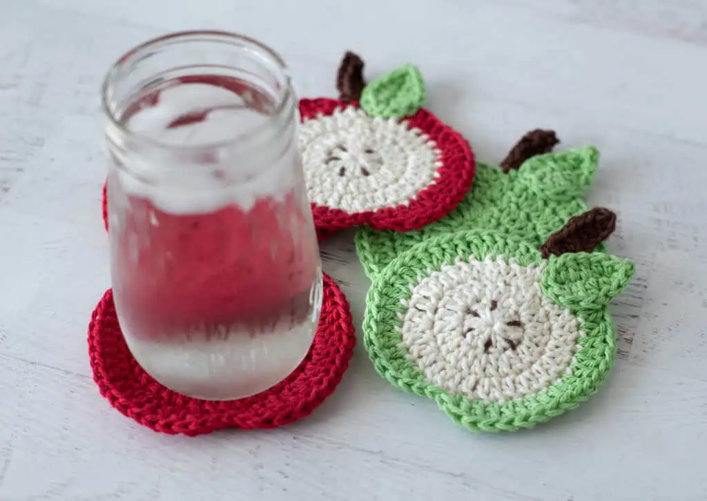Crochet red apple coasters with glass of water. One coaster is a slide of apple with seeds, the rest are red.