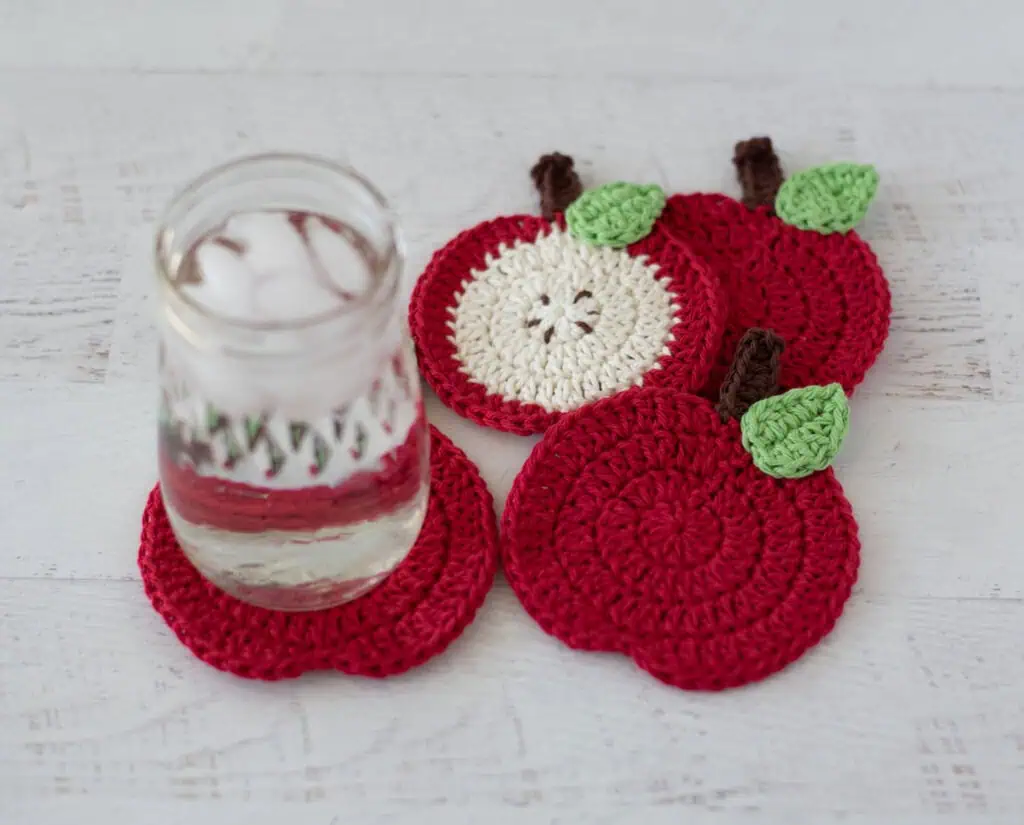 Crochet red apple coasters with glass of water. One coaster is a slide of apple with seeds, the rest are red.