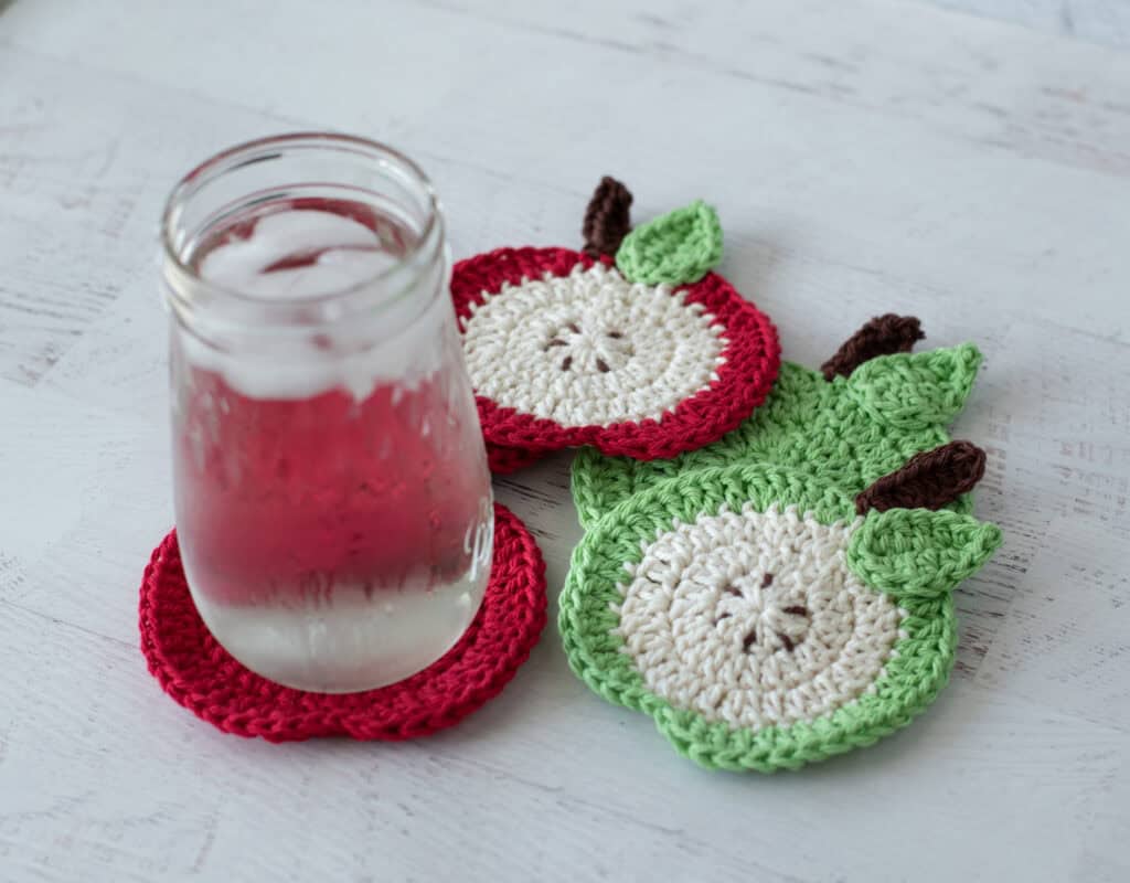 Crochet red apple coasters with glass of water. One coaster is a slide of apple with seeds, the rest are red.