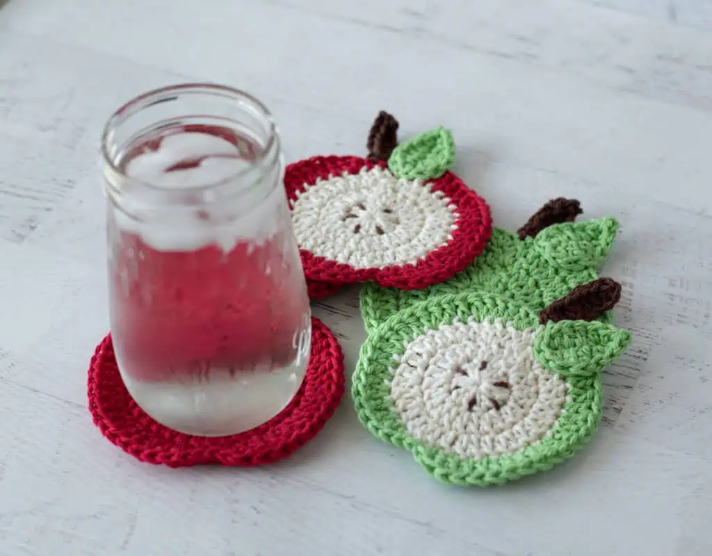 Crochet red apple coasters with glass of water. One coaster is a slide of apple with seeds, the rest are red.