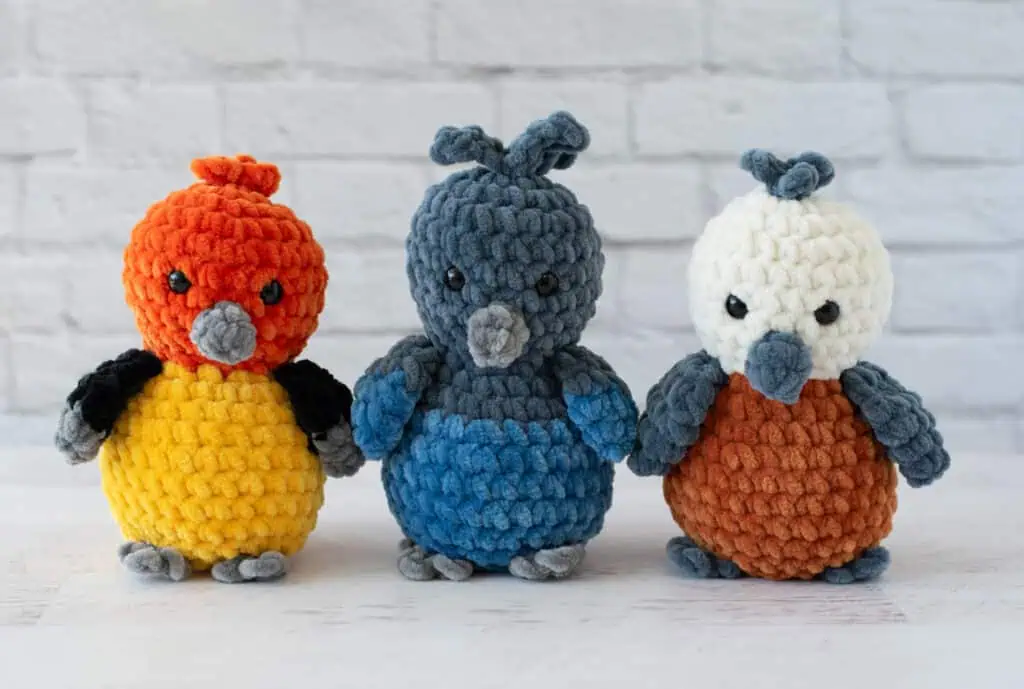3 Crochet birds on a whitewashed wood background with white brick background. Tanager bird is orange, gray, black and yellow. Steller's Jay is dark gray and blue, Nuthatch is white gray and rust.